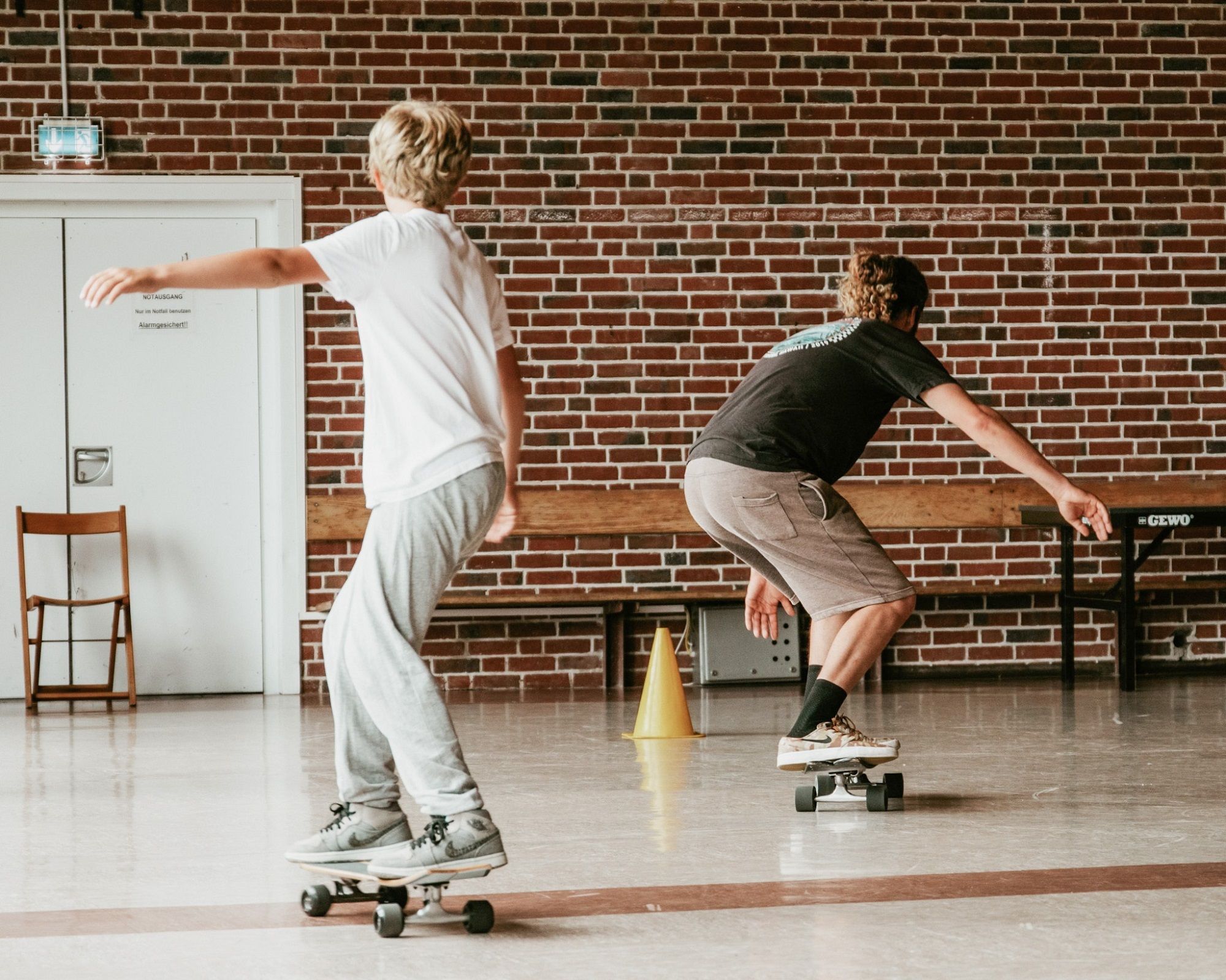 Ein Kind und ein Erwachsener fahren gemeinsam auf Skateboards durch eine Turnhalle mi gemauerter Ziegelwand. Beide befinden sich in einer gebeugten, balancierenden Haltung. Zwischen ihnen steht ein gelber Markierungskegel auf dem Boden. Im Hintergrund sind eine Holzbank, ein Tisch und eine Tür mir einem Notausgangsschild zu sehen.