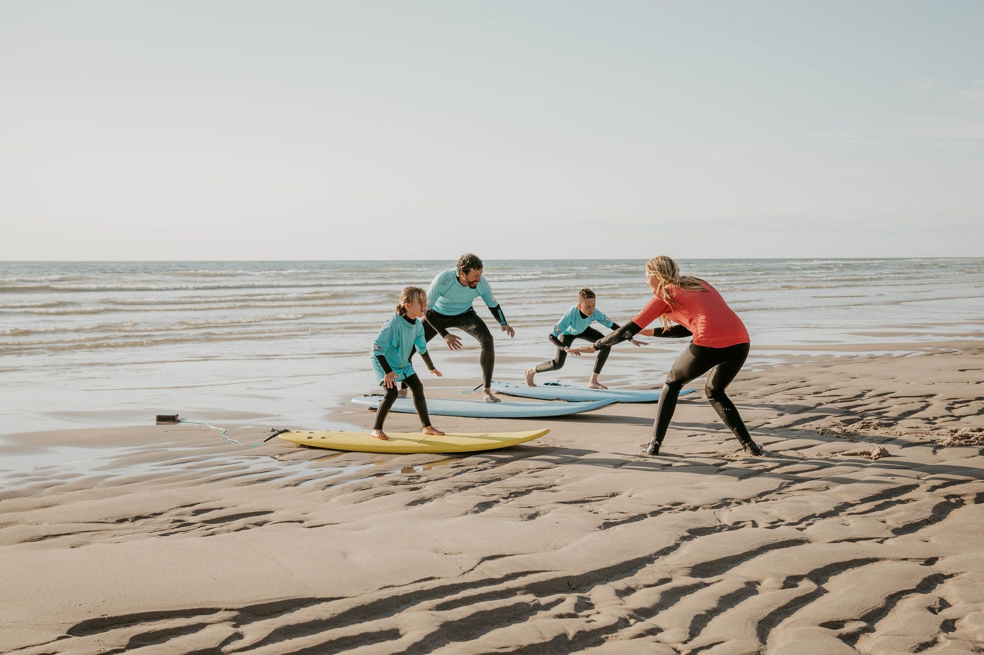 Vier Personen, darunter zwei Kinder und zwei Erwachsene, üben auf dem Sand das Aufstehen auf einem Surfbrett. Eine Surflehrerin in rotem Oberteil und schwarzem Neoprenanzug zeigt die Bewegung vor, während die anderen in türkisfarbenen Shirts und Neoprenanzügen ihr folgen. Die Szene spielt bei Ebbe am Strand mit ruhigem Meer im Hintergrund und weichem Abendlicht.