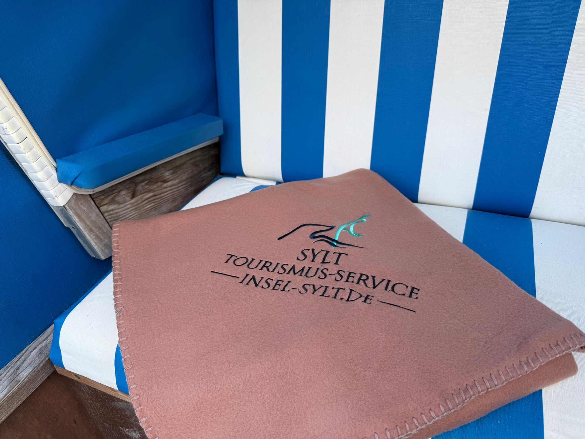 Moccafarbene Strandkorbdecke mit aufgesticktem ISTS-Logo liegt gefaltet im blau-weiß gestreiften Strandkorb.