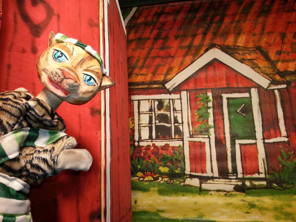 Eine Katzenfigur namens Findus steht vor einem Bild von einem roten Holzhaus.