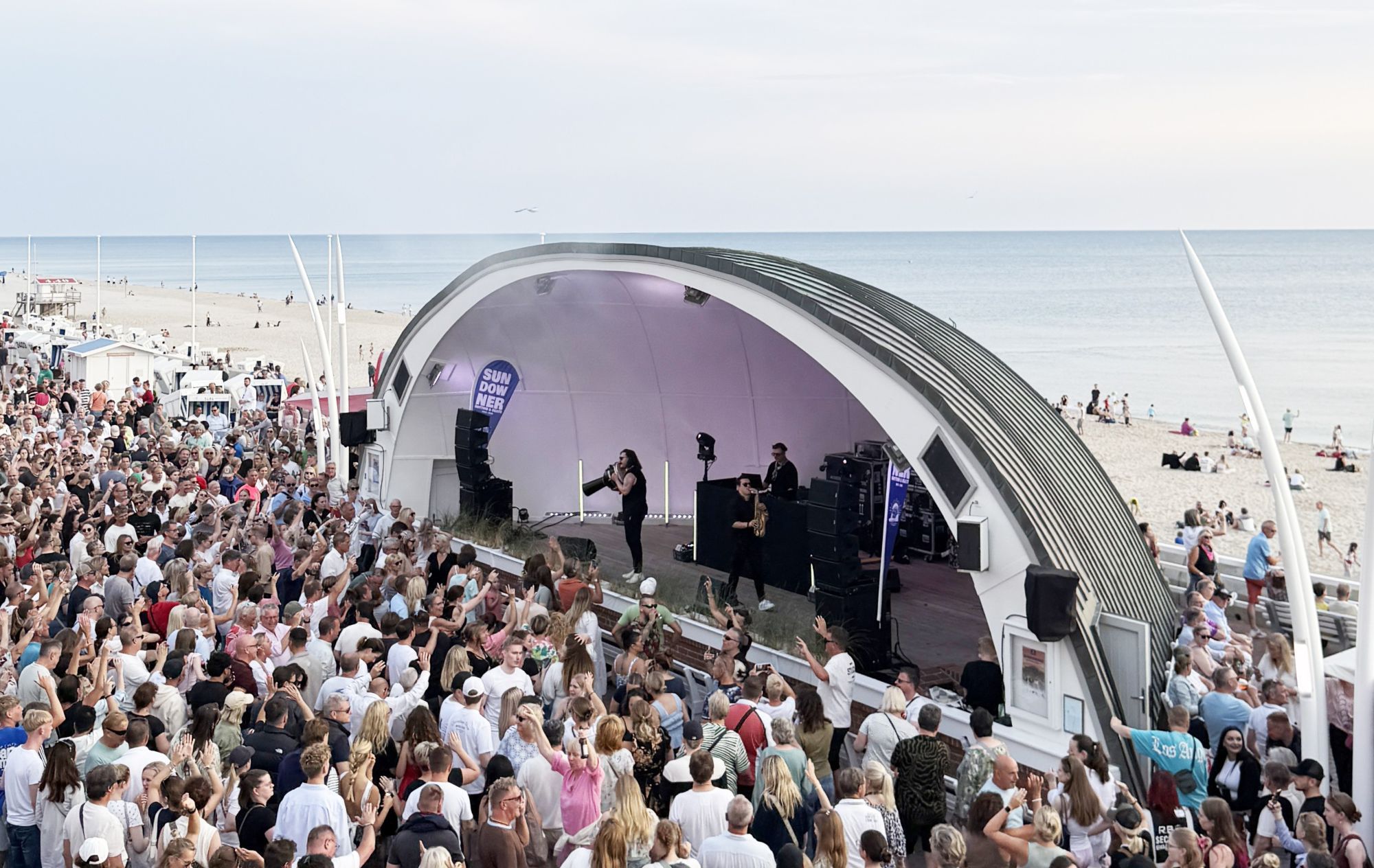 DJ N-Sure, Valerio Lombardo und Luqo beim Sundowner Rhythm & Beats in der Musikmuschel in Westerland auf Sylt, davor eine große Menge feiernde Menschen, dahinter die Nordsee