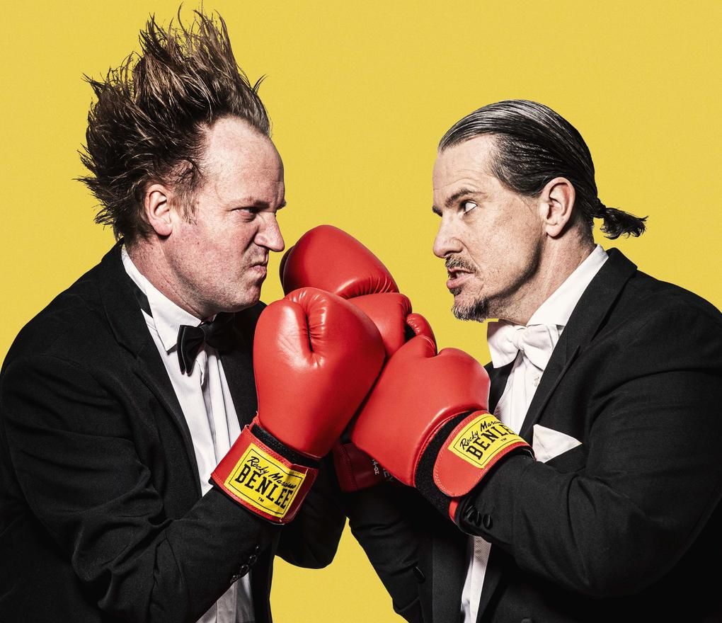 Zwei Männer mit roten Boxhandschuhen und im schwarzen Anzug stehen sich gegenüber vor einer gelben Wand