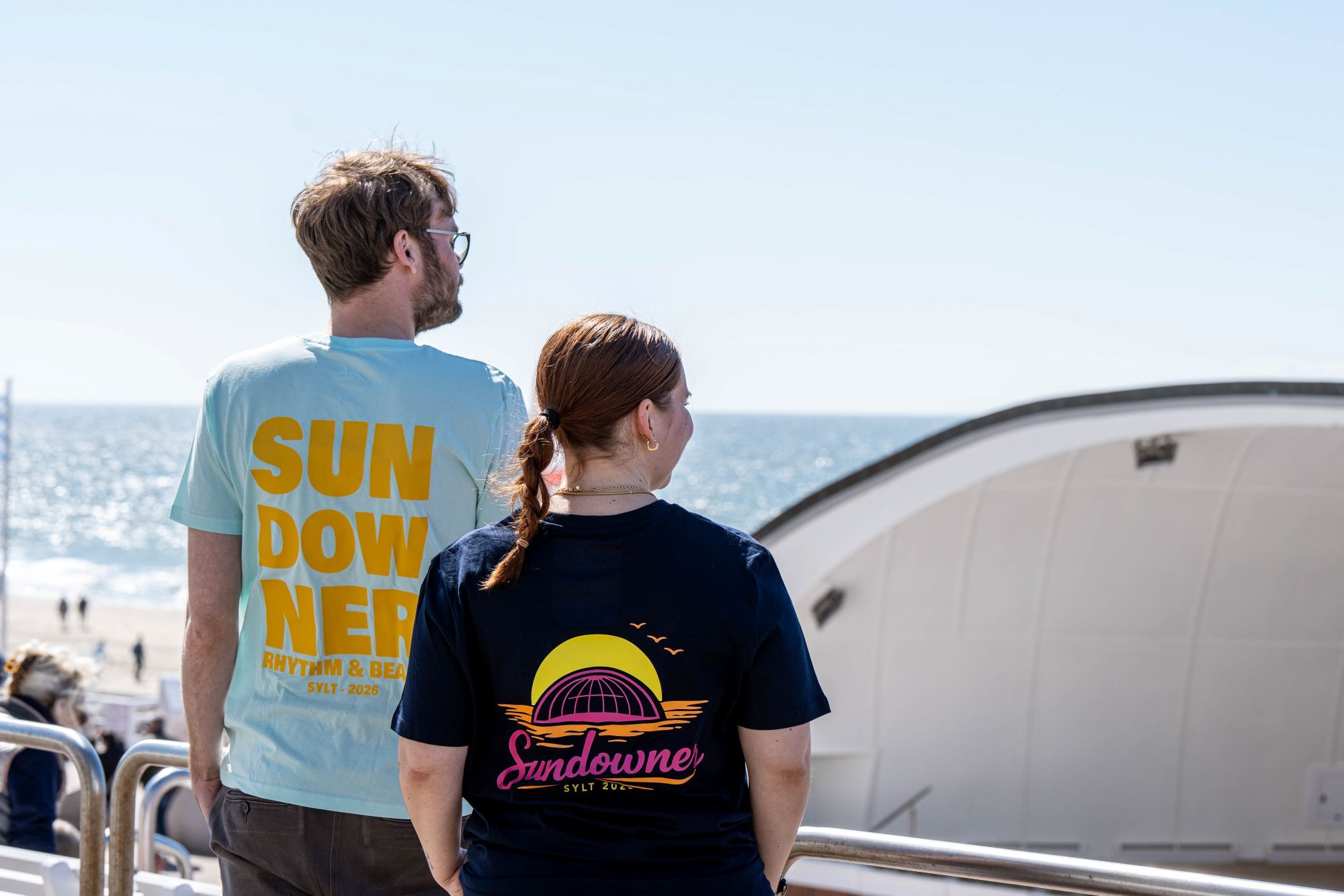 Ein Mann und eine Frau stehen vor der Musikmuschel und tragen die Sundowner T-Shirts 2026