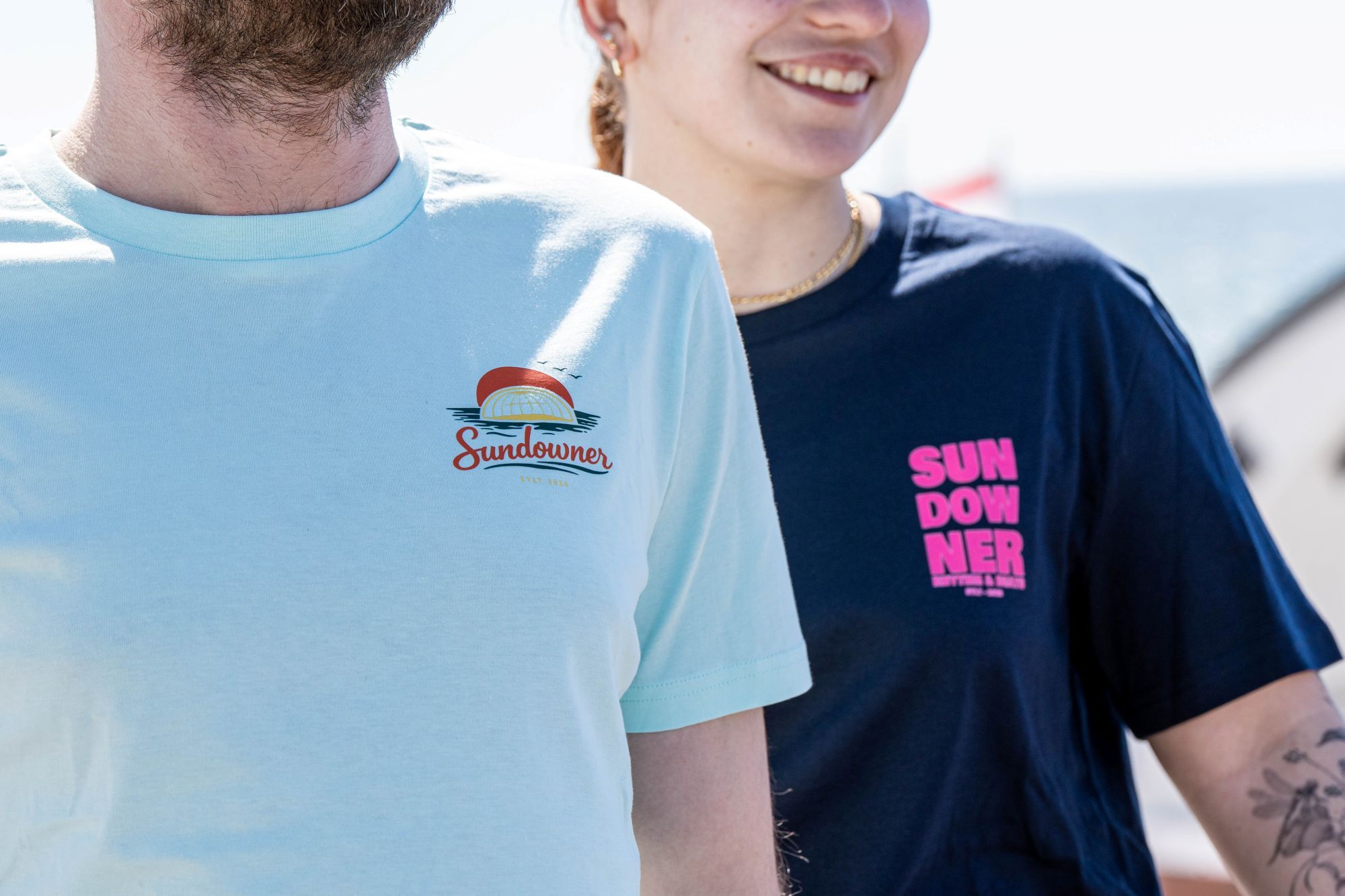 Ein Mann und eine Frau stehen vor der Musikmuschel und tragen die Sundowner T-Shirts 2026