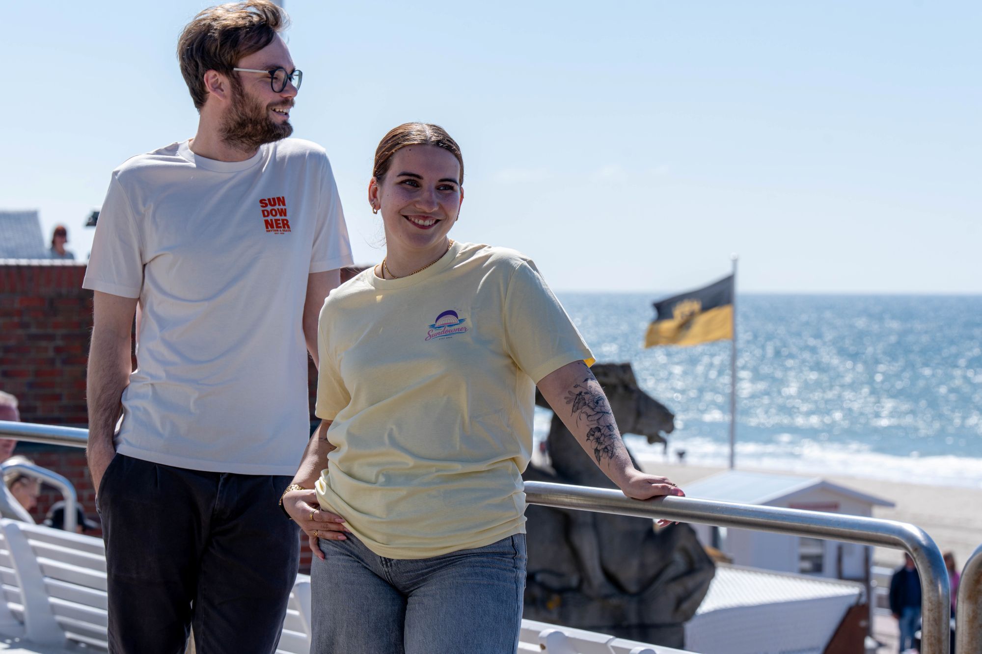 Ein Mann und eine Frau stehen vor der Musikmuschel und tragen die Sundowner T-Shirts 2026