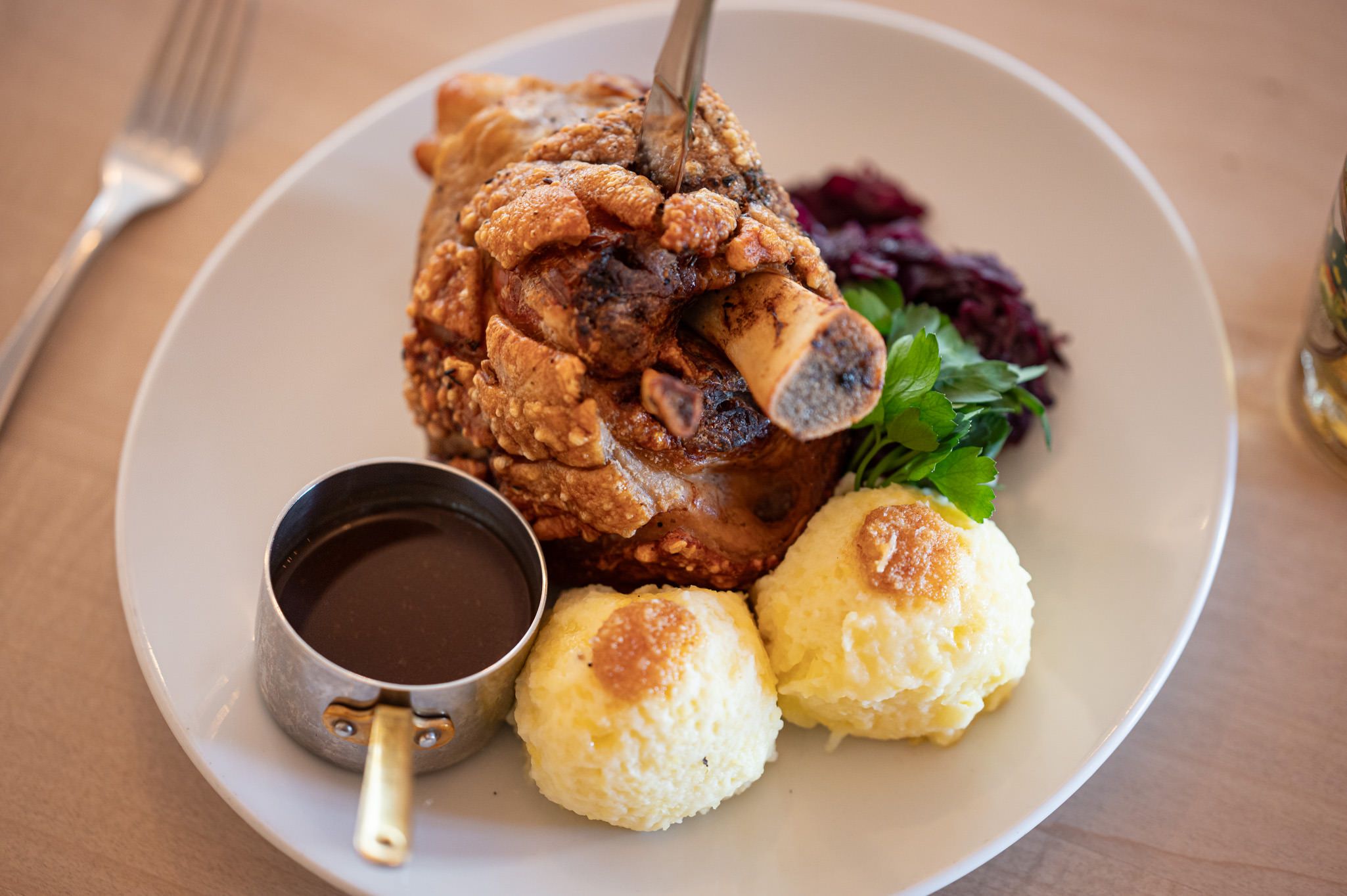 Knusprige Schweinshaxe mit zwei Kartoffelknödeln, Bratensoße, Rotkraut und frischer Petersilie. Ein Klassiker der bayerischen Küche, liebevoll serviert.