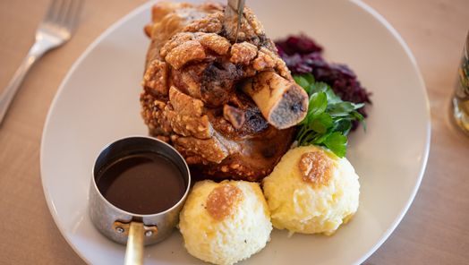 Knusprige Schweinshaxe mit zwei Kartoffelknödeln, Bratensoße, Rotkraut und frischer Petersilie. Ein Klassiker der bayerischen Küche, liebevoll serviert.