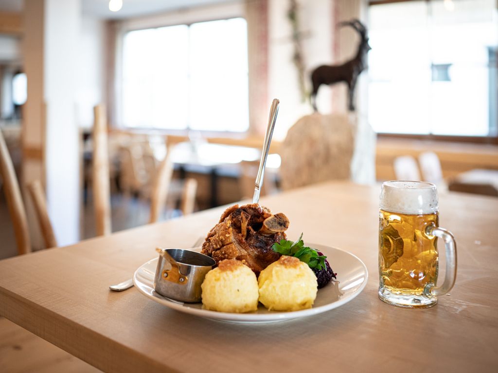 Herzhaft angerichteter Teller mit Knödeln und Schweinehaxe und einem Glas Bier auf einem Tisch im Vordergrund.