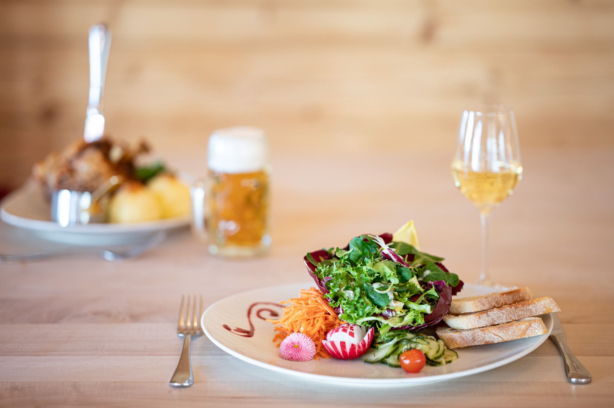 Frischer gemischter Salat mit Brot im Vordergrund, im Hintergrund in unscharf: Haxe mit Knödeln und ein Bierglas.