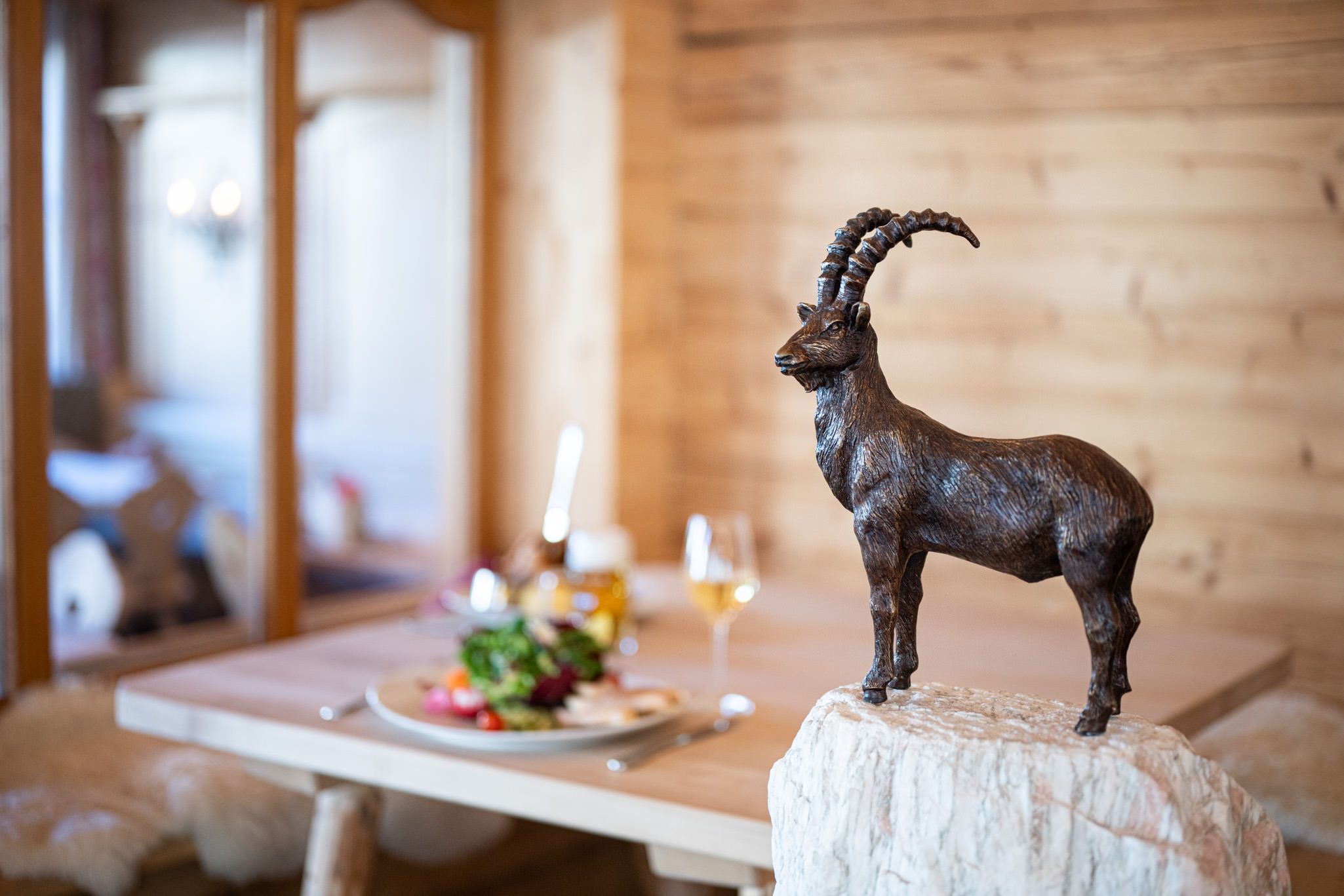 Steinbockfigur im Vordergrund, dahinter ein gedeckter Holztisch mit Salat und Getränken.