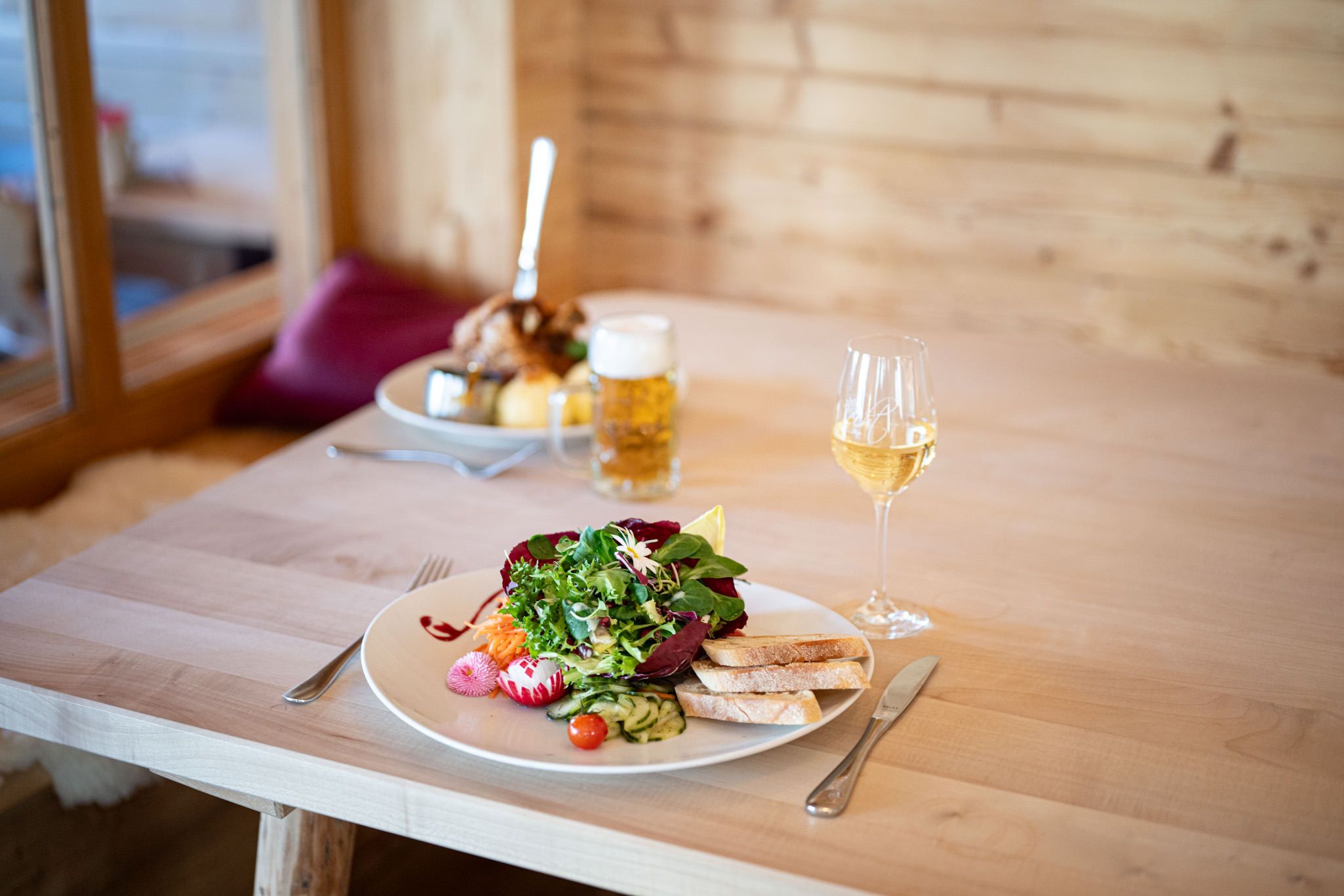 Frischer gemischter Salat mit Brot, dazu ein Glas Weißwein. Im Hintergrund ein Teller mit Haxe, Knödeln und Bier.