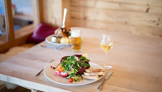 Frischer gemischter Salat mit Brot, dazu ein Glas Weißwein. Im Hintergrund ein Teller mit Haxe, Knödeln und Bier.