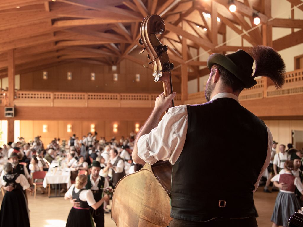 Kontrabassist in Tracht spielt auf Bühne, Gäste tanzen und feiern in festlich geschmücktem Saal.