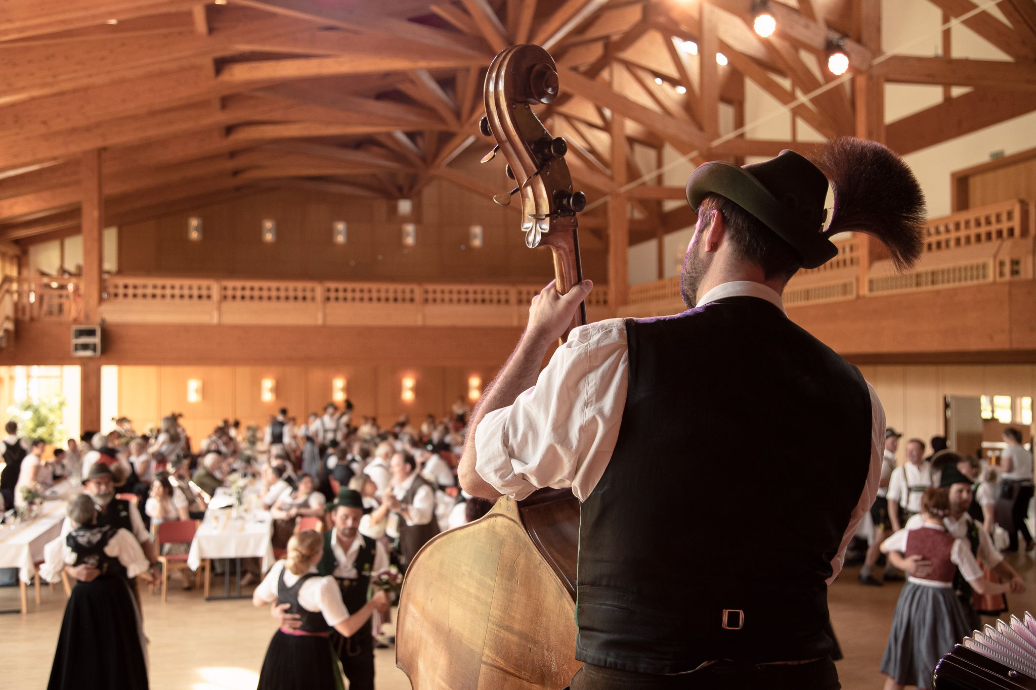 Kontrabassist in Tracht spielt auf Bühne, Gäste tanzen und feiern in festlich geschmücktem Saal.