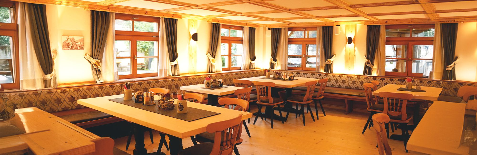 Heller Gastraum mit großen Fenstern, Holztischen und rundum laufender Sitzbank.