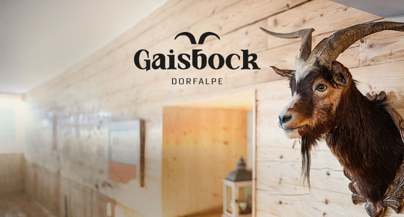 Eine Gaisbock-Trophäe an einer Holzwand unterstreicht das rustikale Ambiente der Dorfalpe, darüber das Gaisbock-Logo als Gestaltungselement.