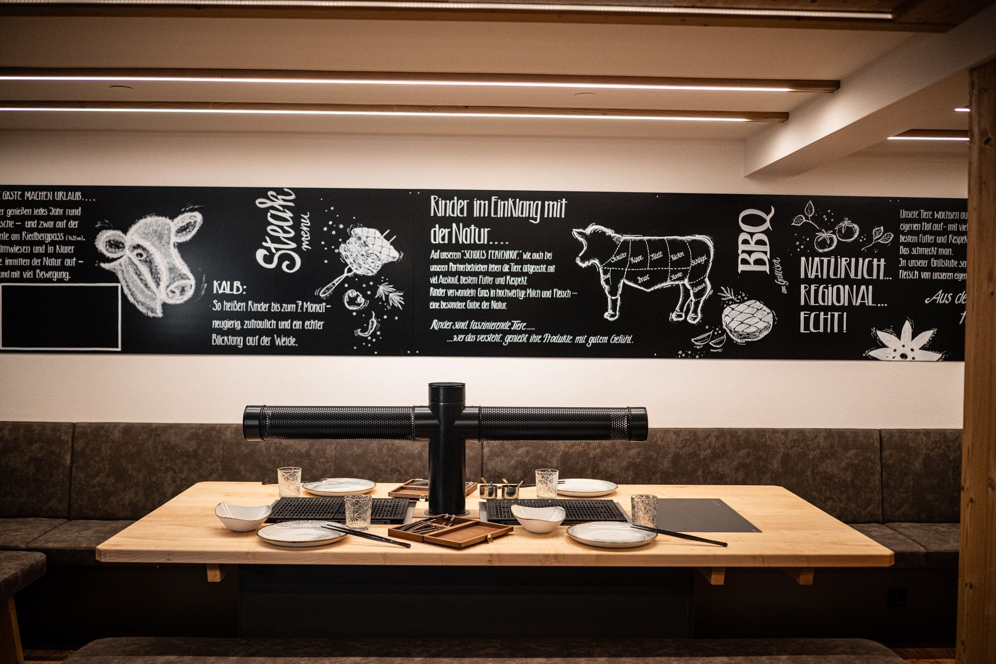 Gemütliche Sitznische in der Grillstube mit großem Holztisch, eingebautem Grill und gedeckten Tellern; an der Wand eine schwarze Tafel mit Illustrationen und Texten rund ums Thema Steak, BBQ und Regionalität