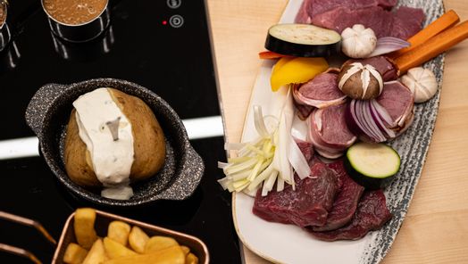 Platte mit frischen Grillzutaten wie Fleisch, Gemüse und Knoblauch, dazu Pommes, Ofenkartoffel und Dips – alles vorbereitet für den Tischgrill in der Grillstube.