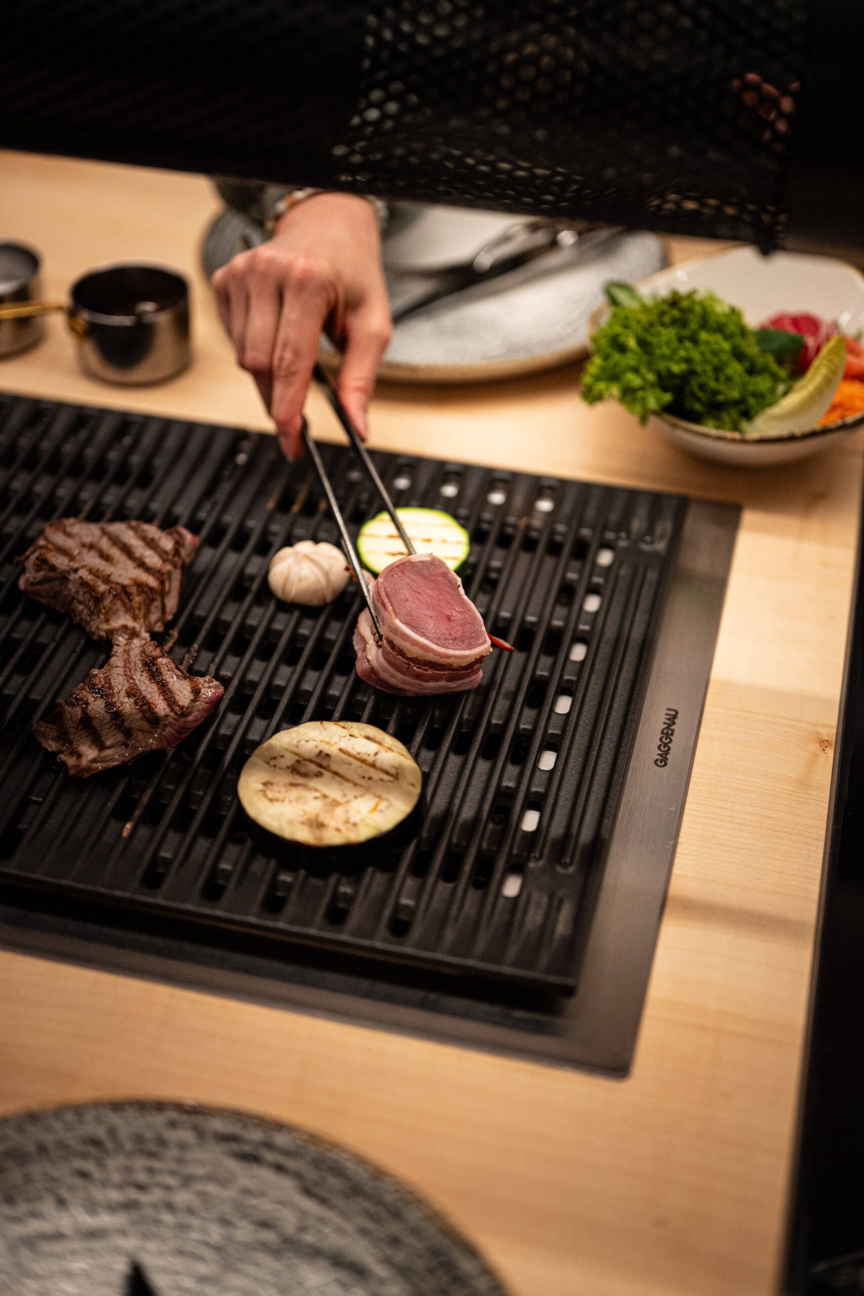 Nahaufnahme beim Tischgrillen: eine Hand legt zart rosa Fleisch und Gemüse auf den heißen Grill; im Hintergrund Schälchen mit frischem Salat und Beilagen – stimmungsvolles Grillvergnügen in der Grillstube.