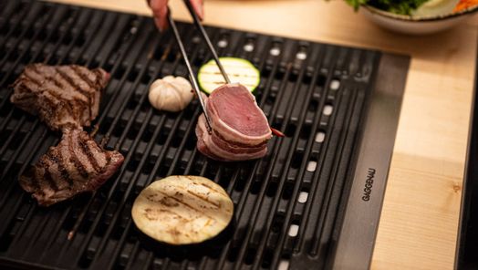 Nahaufnahme beim Tischgrillen: eine Hand legt zart rosa Fleisch und Gemüse auf den heißen Grill; im Hintergrund Schälchen mit frischem Salat und Beilagen – stimmungsvolles Grillvergnügen in der Grillstube.