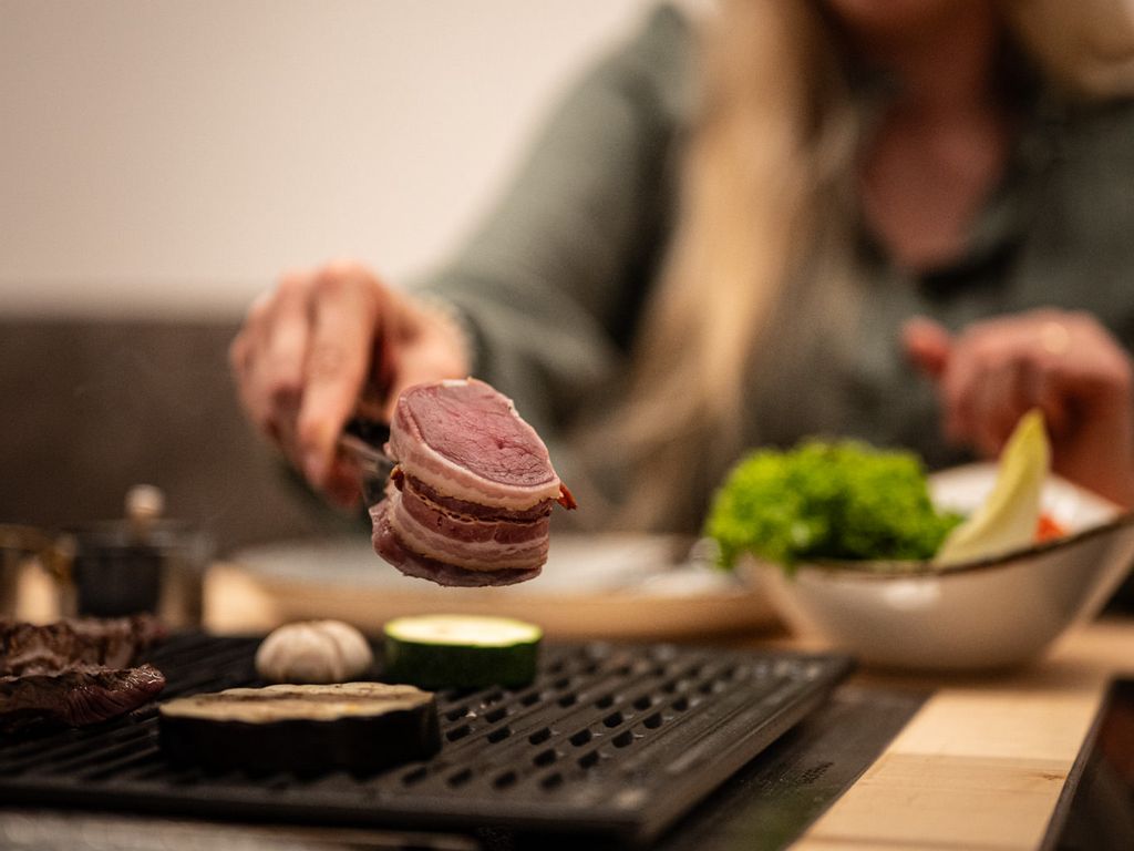 Nahaufnahme beim Tischgrillen: eine Person legt ein Stück Fleisch auf den heißen Grill, daneben brutzeln Gemüse und Knoblauch – gemütliche Atmosphäre in der Grillstube.