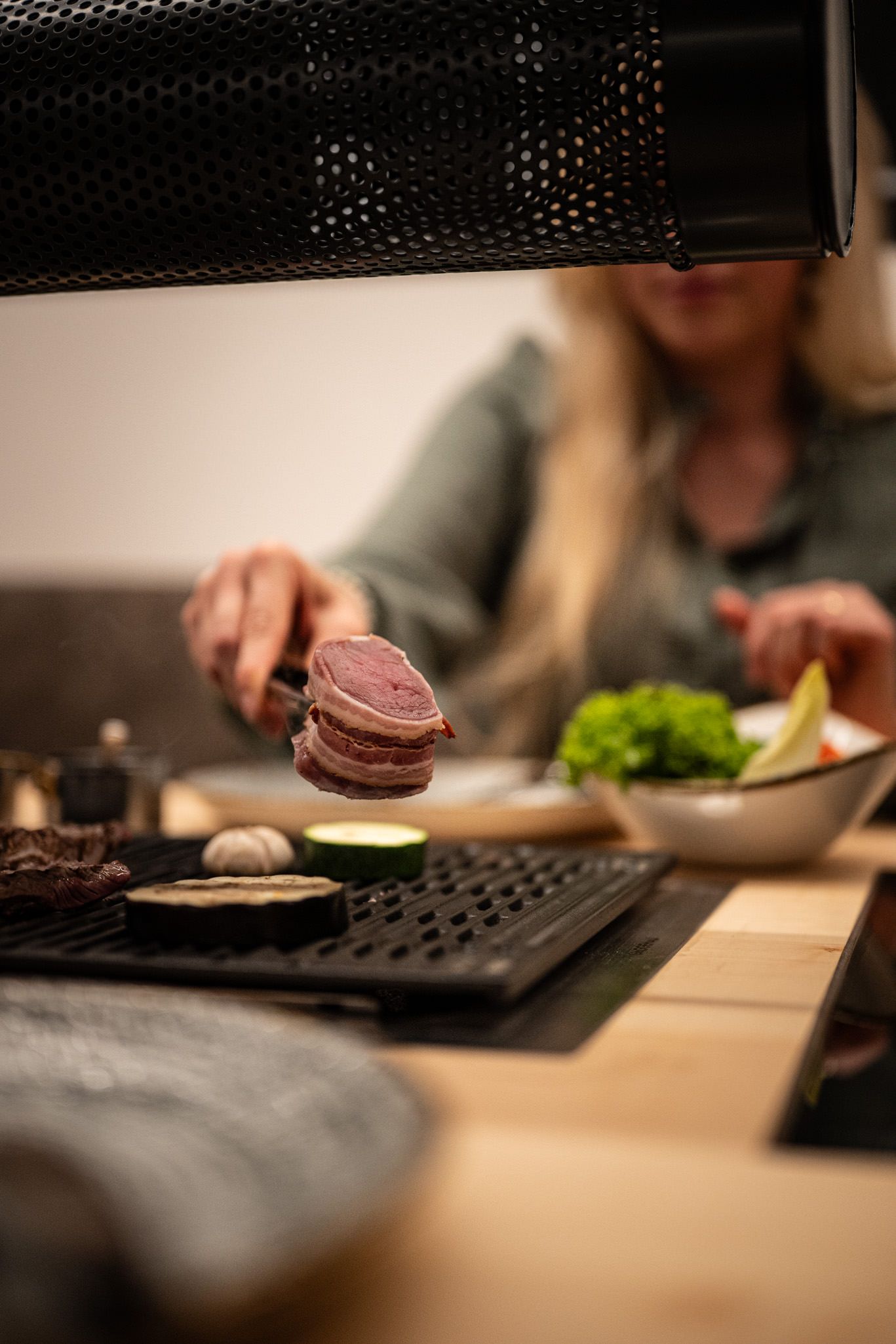 Nahaufnahme beim Tischgrillen: eine Person legt ein Stück Fleisch auf den heißen Grill, daneben brutzeln Gemüse und Knoblauch – gemütliche Atmosphäre in der Grillstube.