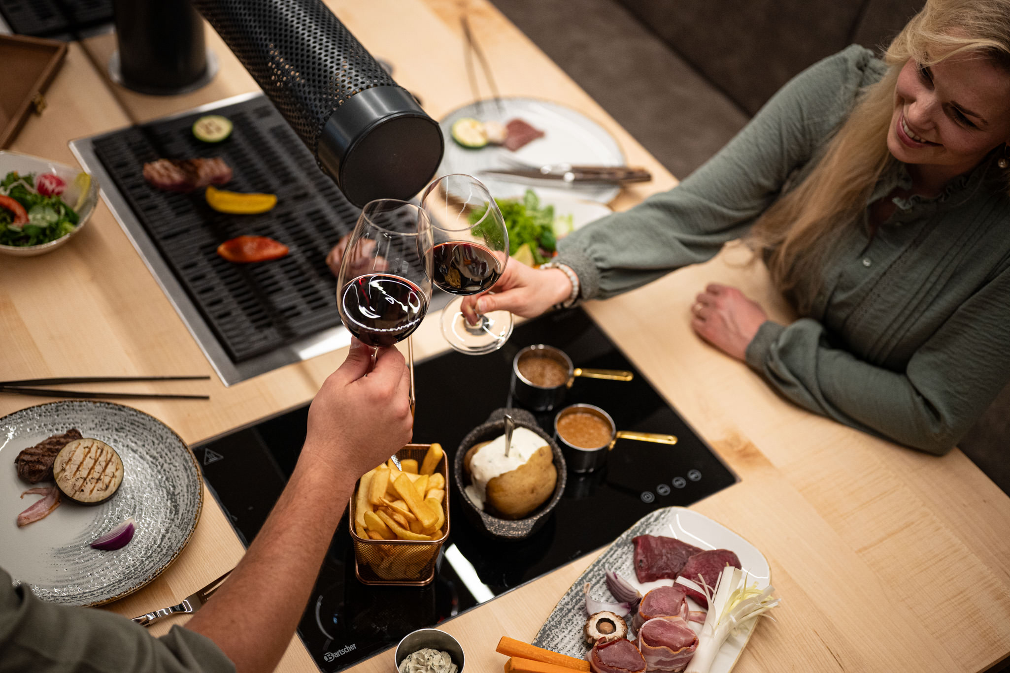 Gesellige Szene am Tischgrill: Zwei Personen stoßen mit Rotwein an, auf dem Grill liegen Gemüse und Fleisch; auf dem Tisch stehen Saucen, Ofenkartoffel, Pommes und eine Platte mit frischen Grillzutaten