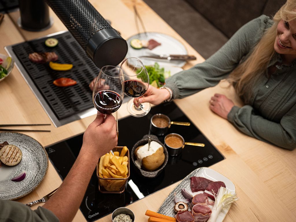 Gesellige Szene am Tischgrill: Zwei Personen stoßen mit Rotwein an, auf dem Grill liegen Gemüse und Fleisch; auf dem Tisch stehen Saucen, Ofenkartoffel, Pommes und eine Platte mit frischen Grillzutaten