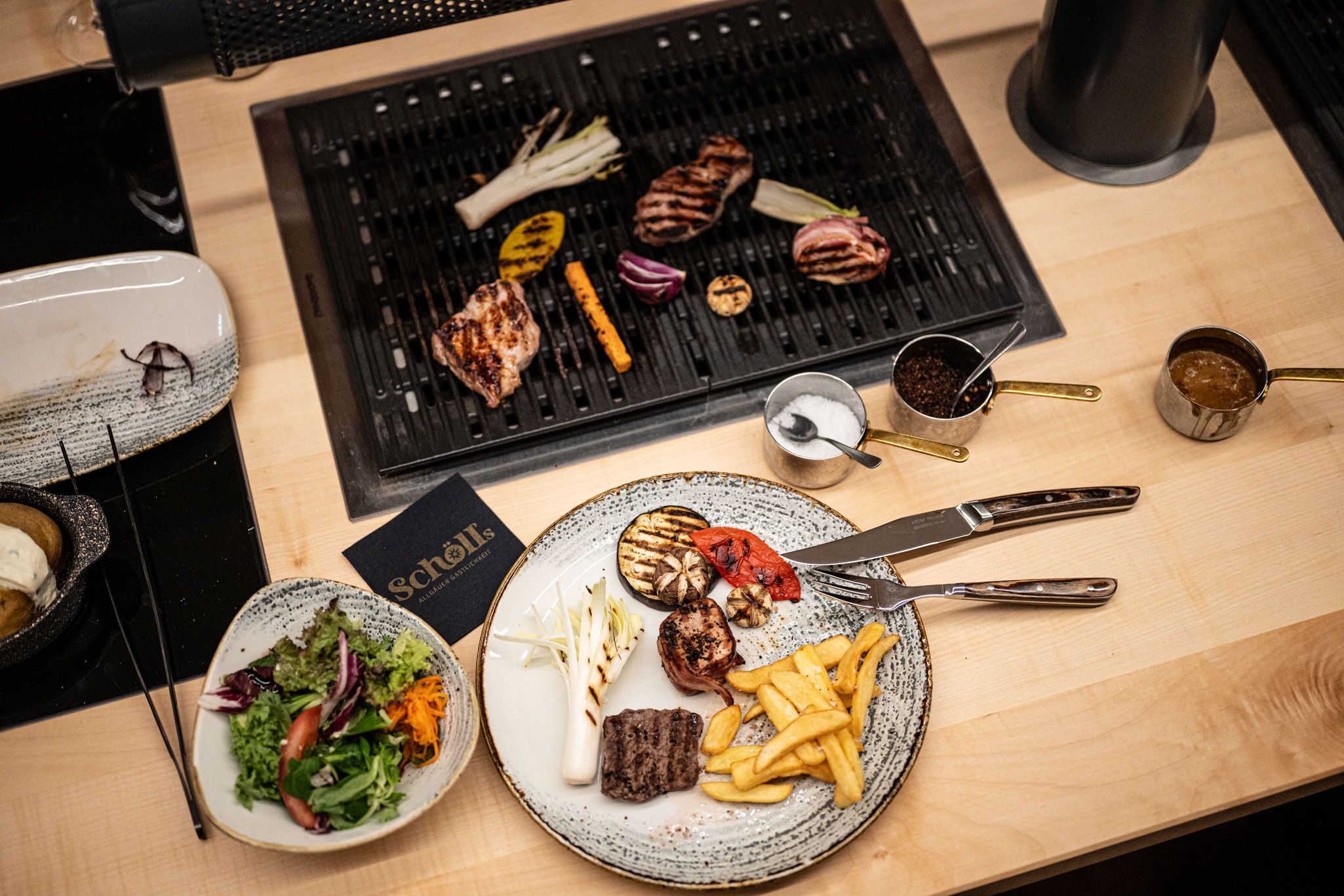 Tischgrill mit gegrilltem Fleisch und Gemüse, dazu ein Teller mit Pommes, Salat und Dips – ein genussvoll angerichtetes Grillgericht in gemütlicher Atmosphäre